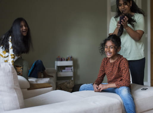 Två flickor sitter i sängen och en vuxen kvinna borstar den enas hår. 