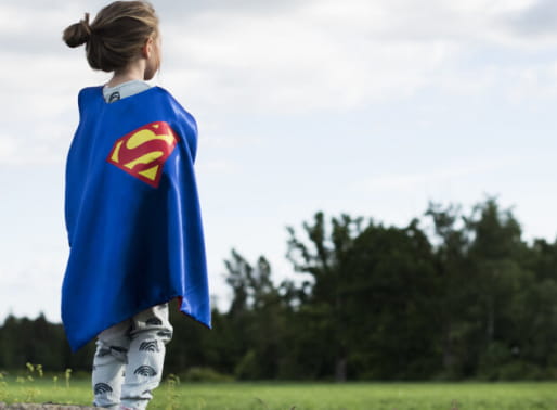 Barn med superhjälte-mantel står med ryggen mot och ser ut över en äng.