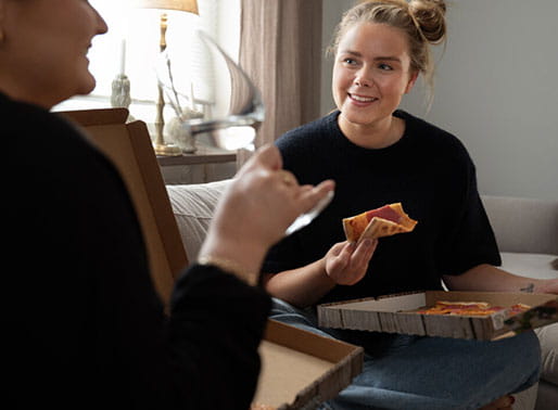 Två glada kvinnor äter pizza i soffan.