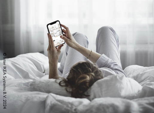 En tjej ligger på rygg i en säng med vita sängkläder och tittar på sin mobil.